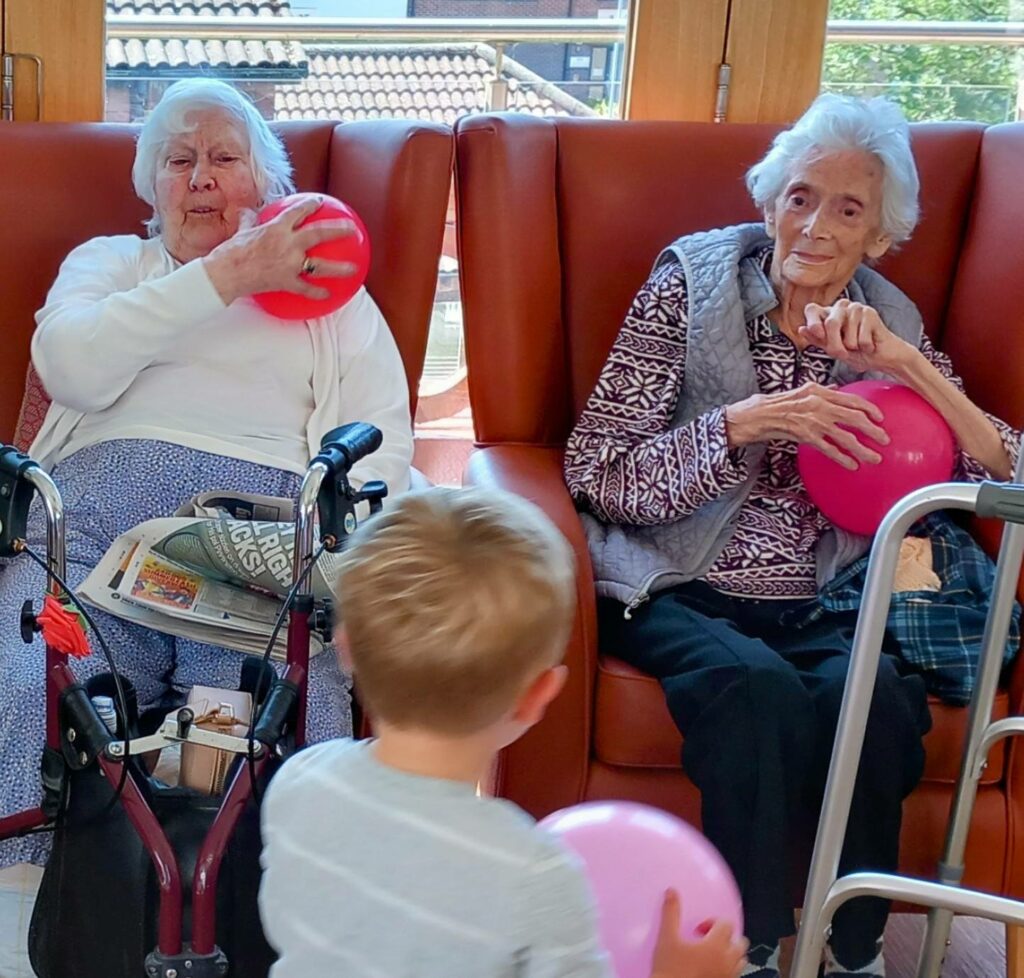 During a joyful visit to St Agnes Pre-School, two elderly women in armchairs hold balloons while a young child stands in front of them with a pink balloon. Walking frames rest beside the women.