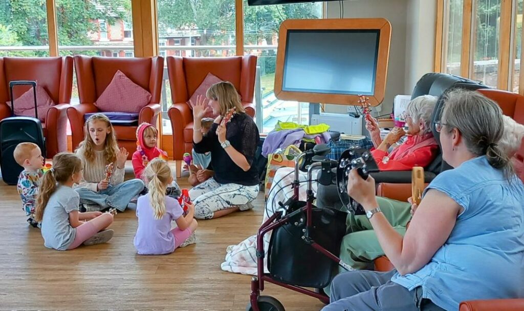A group of children and adults are sitting in a circle on the floor of a bright room; some are holding musical instruments, while others are seated on chairs, either observing or playing music.