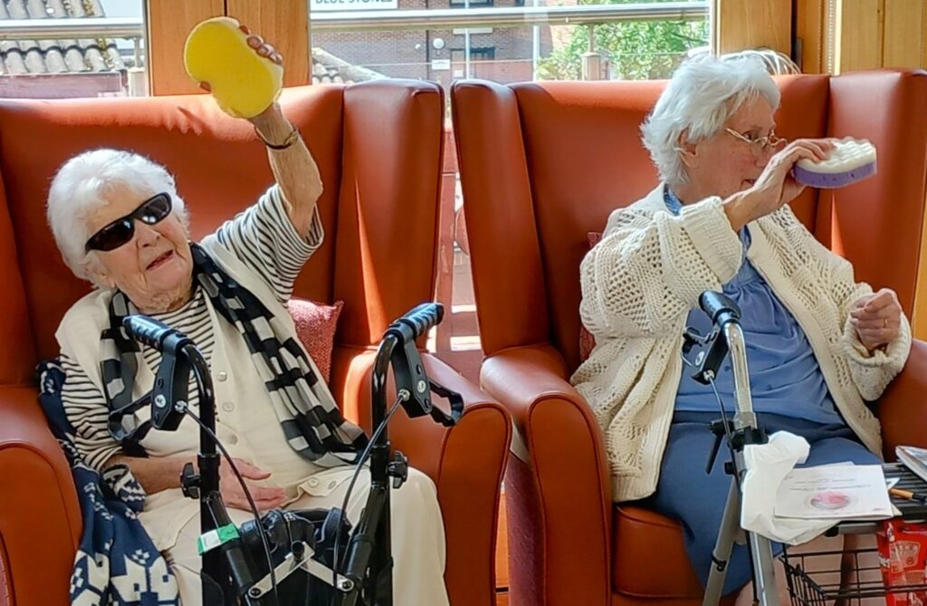 Two elderly women are seated in armchairs, each holding up a sponge while taking part in an activity. Walking frames are visible in front of them.