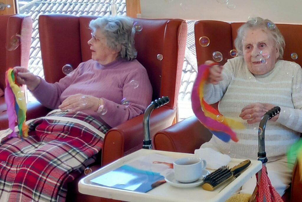 Two elderly women sit in armchairs, waving colourful scarves at floating bubbles in a joyful send-off; a table with a cup and brush sits before them.