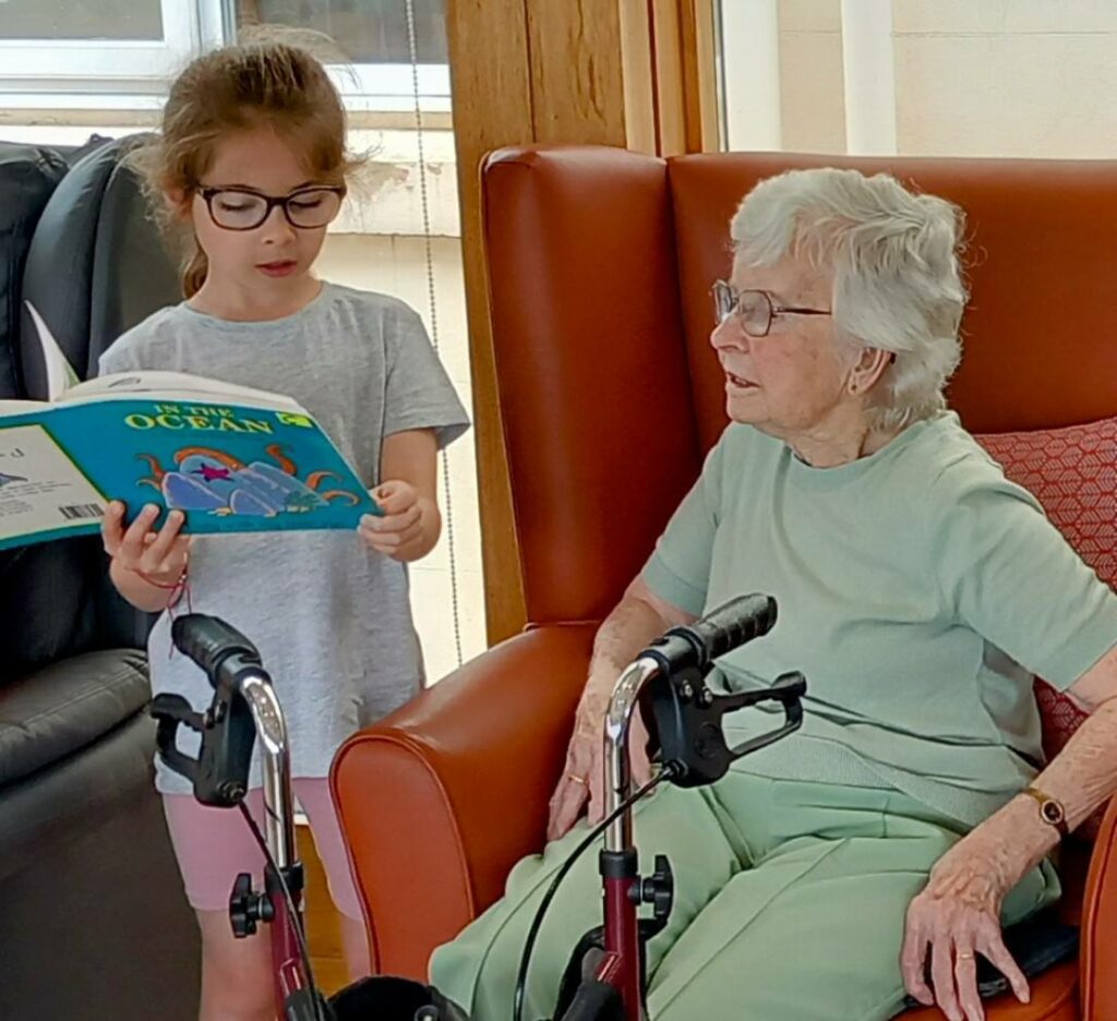 A young girl stands and reads a book aloud to an elderly woman seated in an armchair, with a walking frame in front of her.