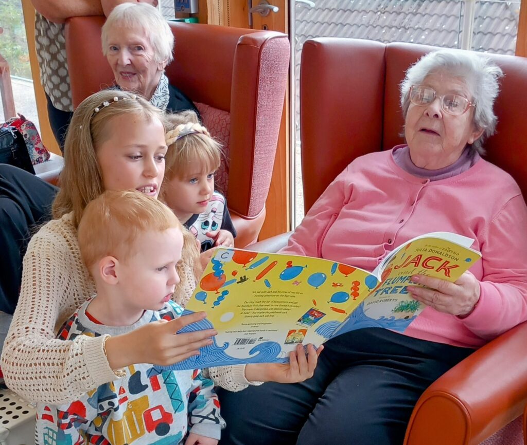 An elderly woman sits in an armchair holding a book while three young children look at it, with another older woman sitting nearby, all indoors.