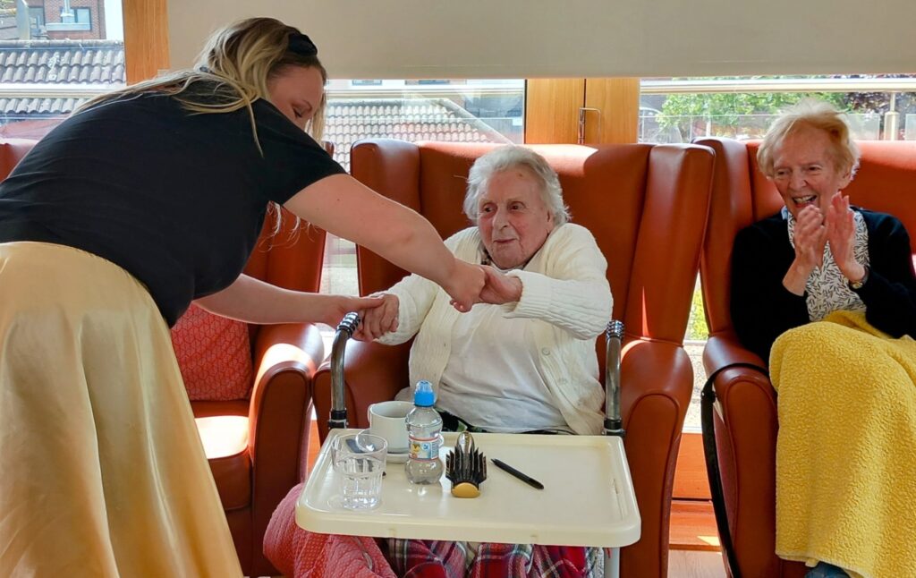 A woman greets an elderly lady seated in a chair with a handshake, while another elderly lady nearby smiles and applauds—creating a joyful scene reminiscent of a kitchen Boogie Woogie Musical Feast. Drinks and nibbles sit on the table.