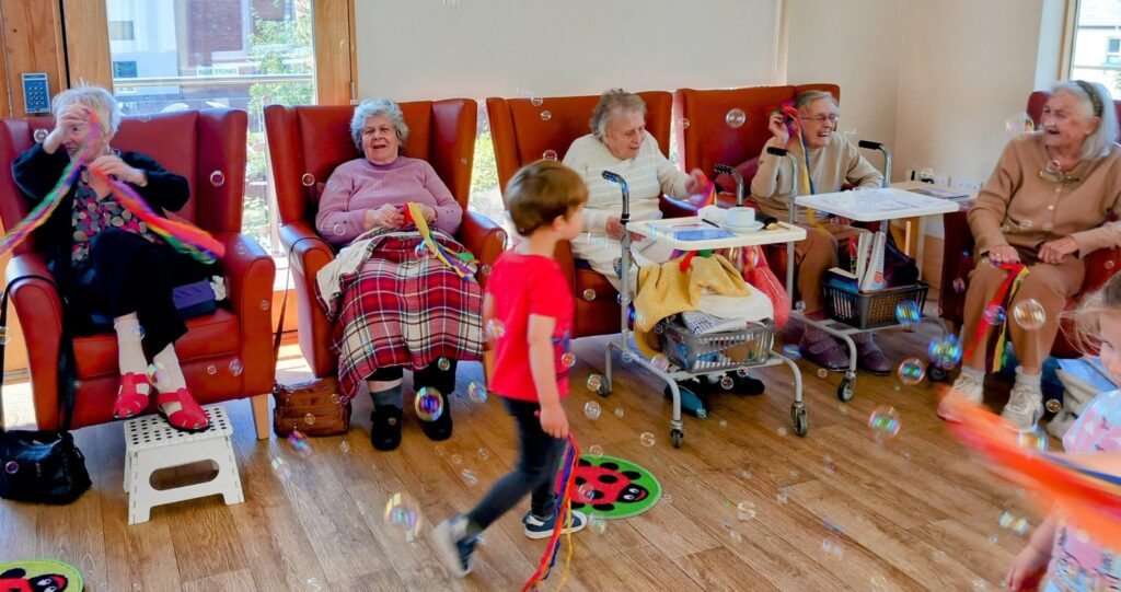A group of elderly women sit in chairs, smiling, while a young child from St Agnes Pre-School walks past them during the final session in a room filled with bubbles.