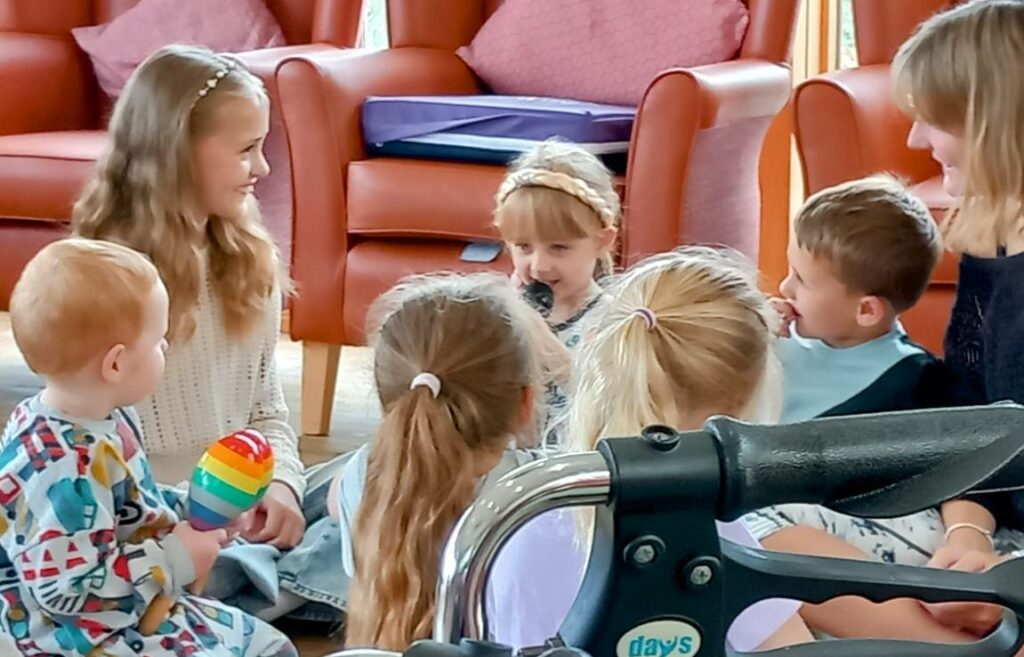 A group of young children are sitting on the floor in a circle with an adult nearby; one child is holding a microphone, and another holds a colourful rattle. A wheelchair is visible in the foreground.
