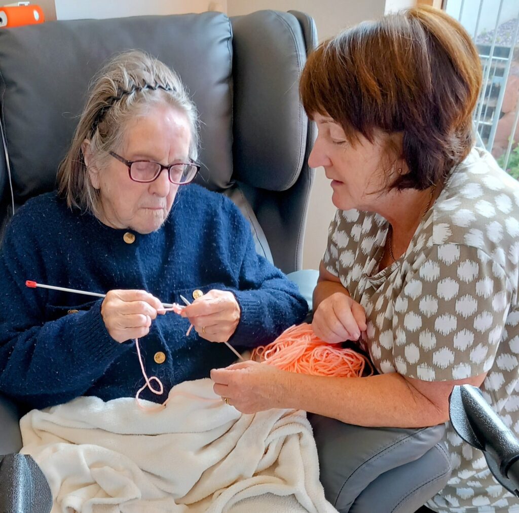 An elderly woman sits in a chair knitting with pink wool, while another woman sits beside her, holding the wool and watching attentively.