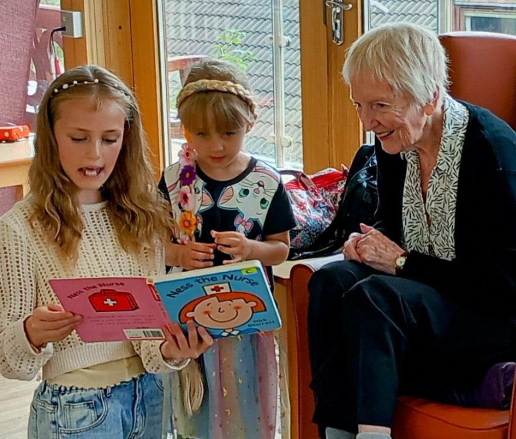 Two young girls are reading books to an elderly woman, who is smiling and listening attentively in a bright room.