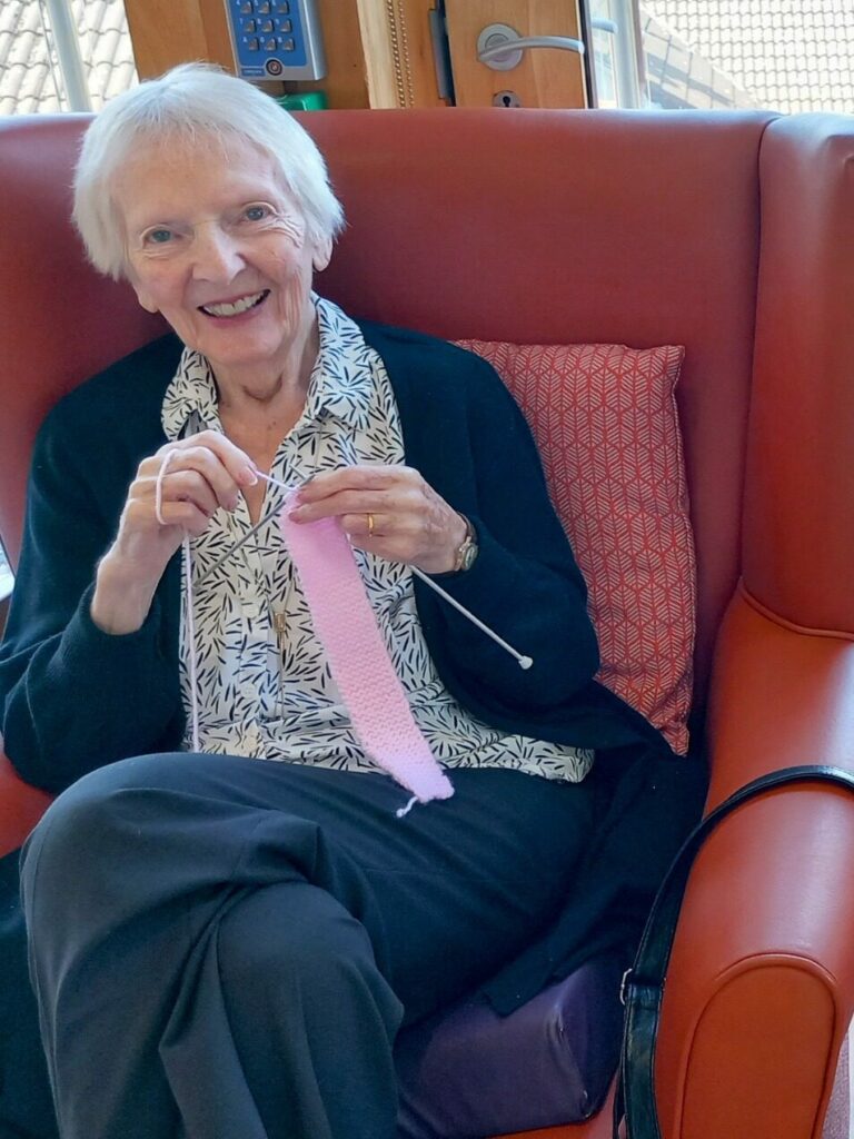 Two elderly women sit in armchairs by a window, smiling and knitting with pink wool and cream-coloured wool.
