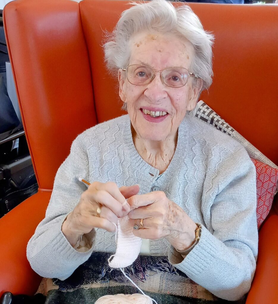 An elderly woman wearing glasses sits in an orange chair, smiling as she crochets with light-coloured yarn.