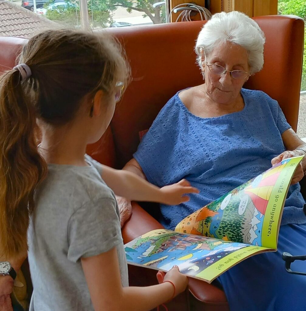 An elderly woman sits in a chair reading a colourful children’s book with a young girl, who is pointing at a page in the book.