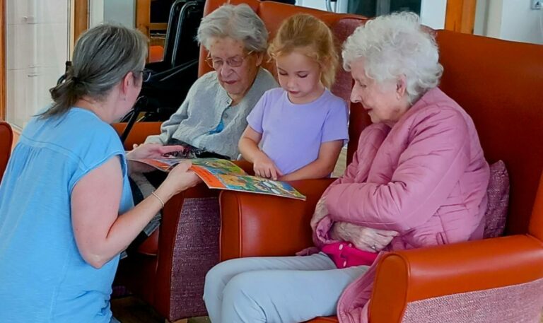 A woman and a young girl show a colourful book to two elderly women seated in armchairs, all engaged and looking at the book.