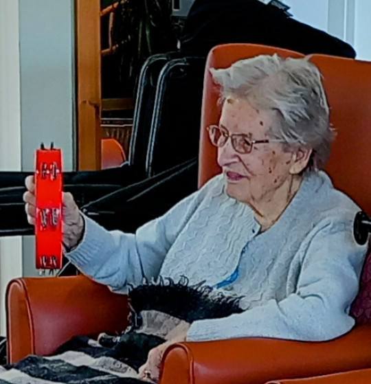 Elderly woman seated in an orange chair, holding a red musical shaker in her right hand, with a blanket on her lap.