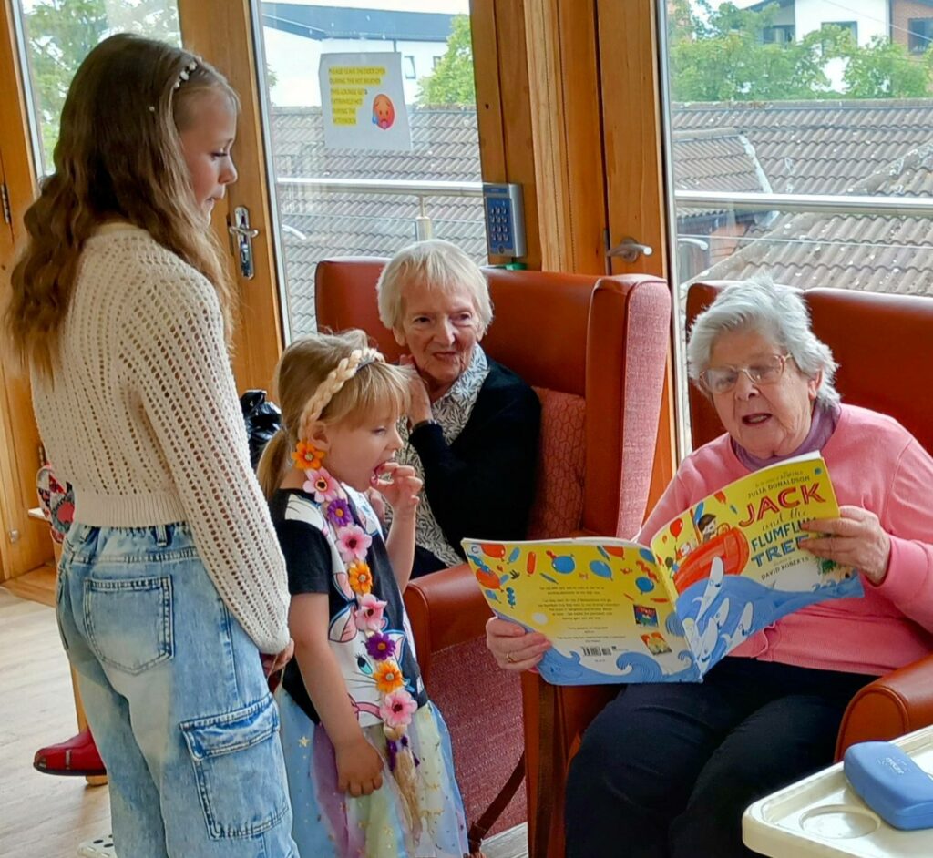 An elderly woman reads a childrens book to two young girls while another elderly woman looks on in a bright room with large windows.