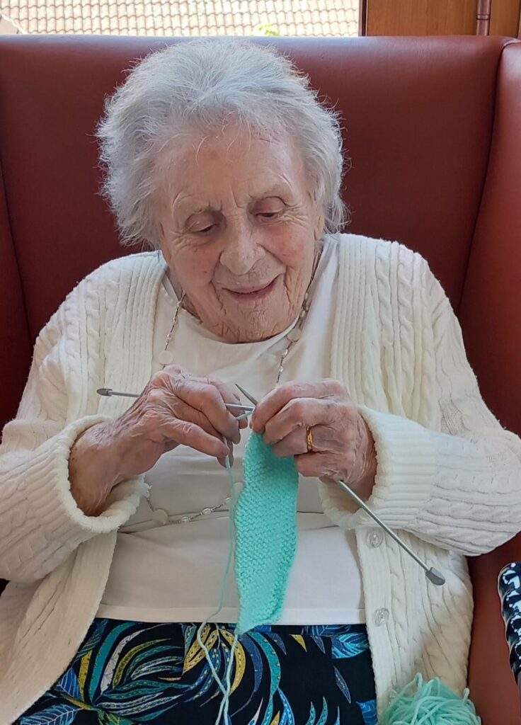An elderly woman with white hair sits in an armchair, knitting a piece of light blue fabric with knitting needles. She is wearing a white cardigan and a patterned skirt.