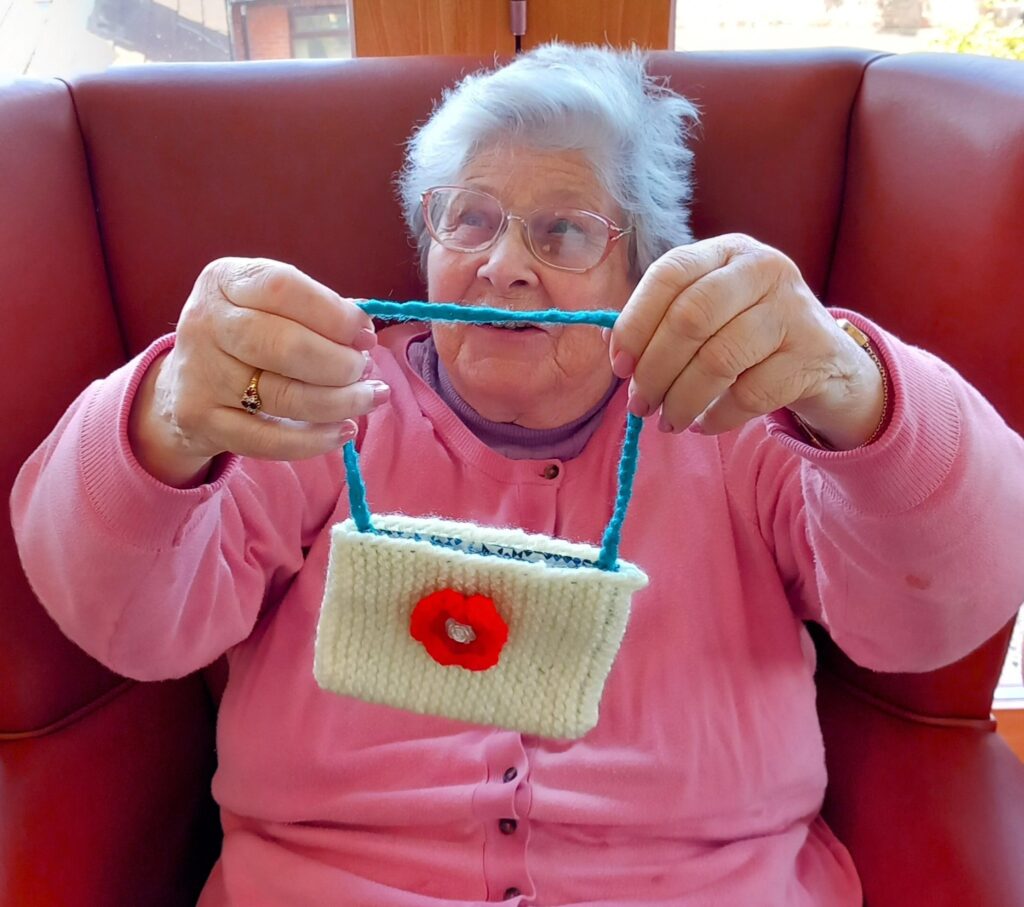 An elderly woman in a pink jumper sits on a red chair, holding up a small, handmade knitted bag with a blue strap and a red flower decoration.