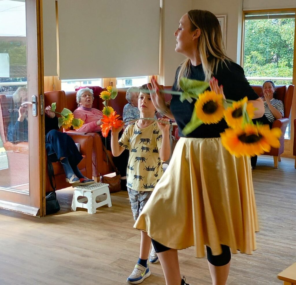 A woman and a child holding artificial sunflowers perform in front of seated elderly people in a lounge with large windows.