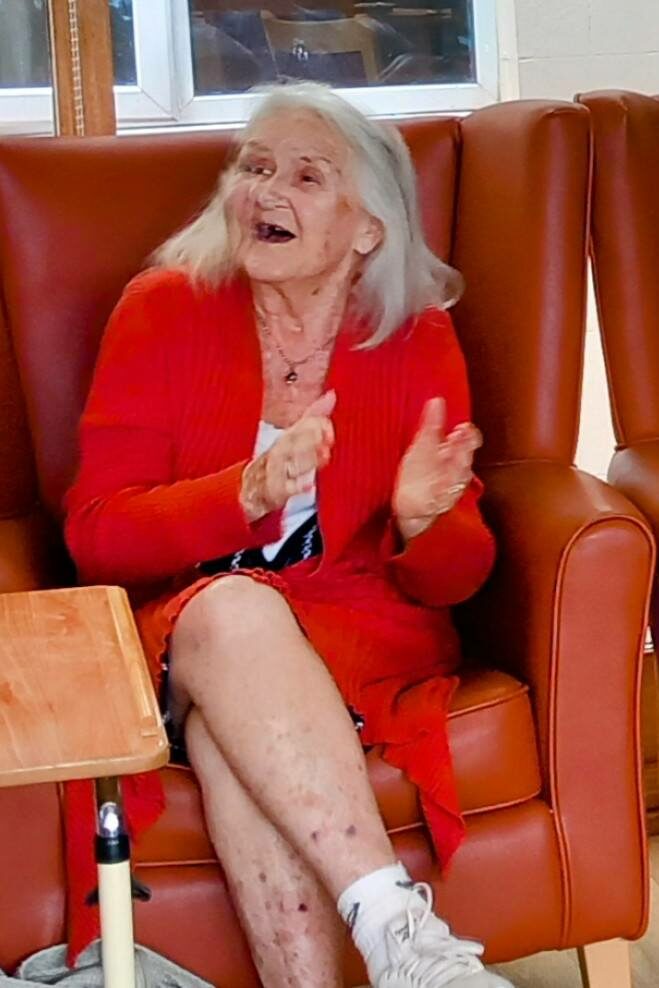 Two elderly women sit in armchairs; one claps her hands while the other plays a red tambourine. Both seem absorbed in the activity, relishing a cheerful afternoon akin to The 64 N’Ukes gatherings.