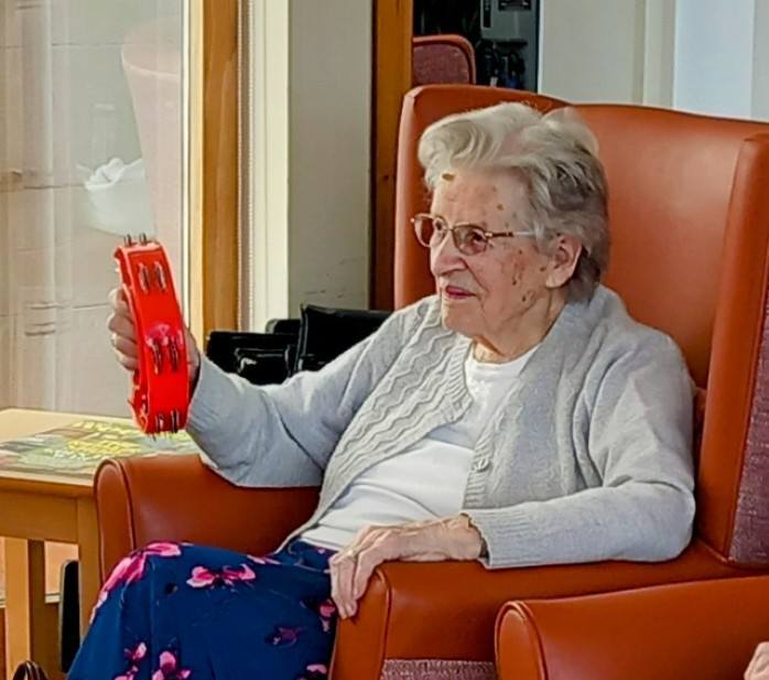 An elderly woman wearing spectacles sits in an armchair, holding a red tambourine in her right hand, ready to join The 64 N’Ukes for a joyful afternoon of music.