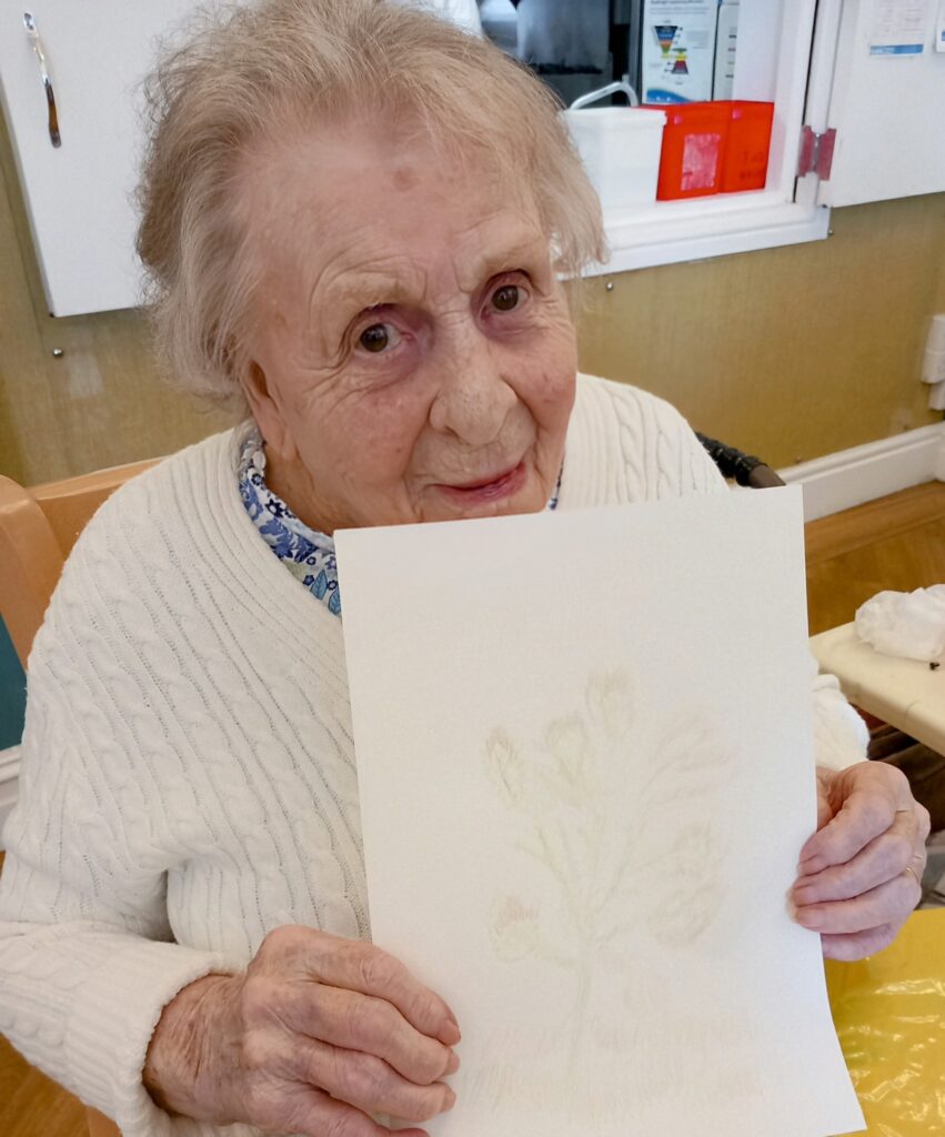 An elderly woman in a white jumper sits indoors, smiling as she holds up a lightly coloured sketch of a tree on a sheet of paper and looks at the camera.