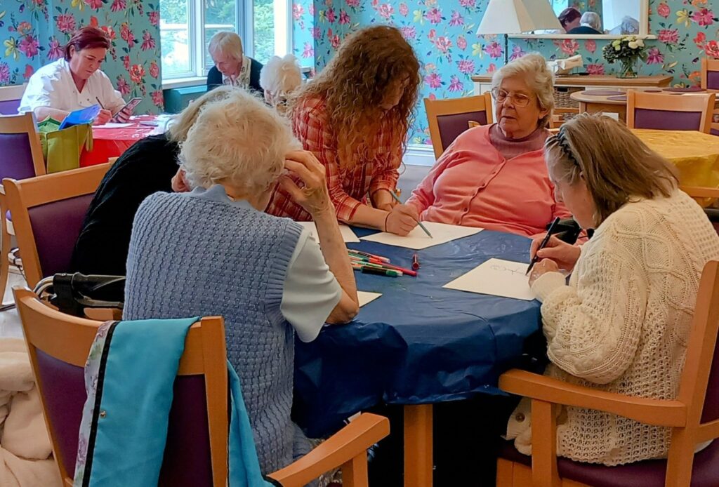 Four elderly women sit smiling at a table, sketching and writing on paper, assisted by a younger woman, in a brightly lit room with floral wallpaper.