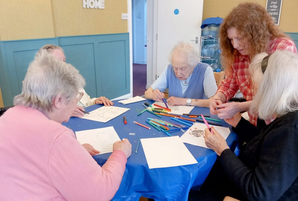 Four elderly women sit around a table, smiling as they sketch and colour with pencils, while a younger woman assists them in a cosy indoor setting.