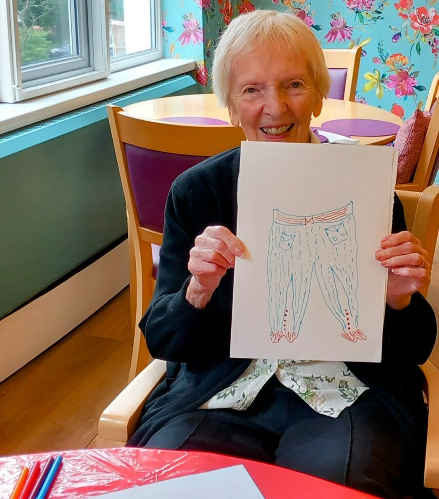 An older woman sits smiling in an armchair, holding up her sketch of a pair of jeans. She is indoors near a window, with colourful floral wallpaper in the background.
