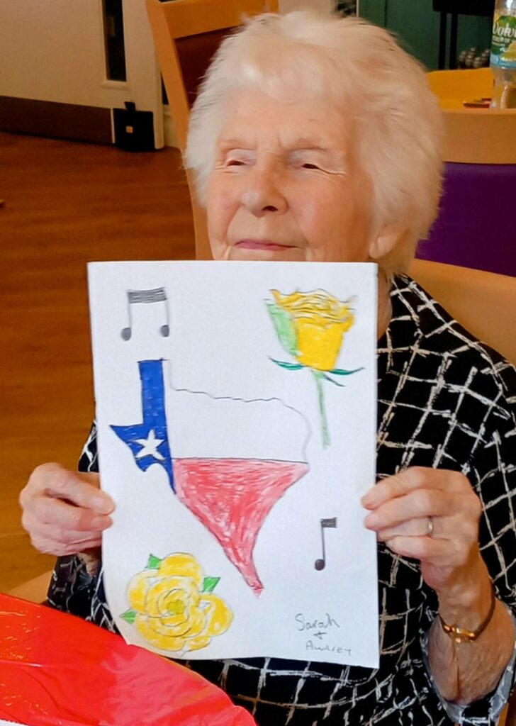 A smiling elderly woman holds up a drawing of Texas adorned with musical notes and yellow roses, sitting indoors at a table, as if sketching her love for music and home.