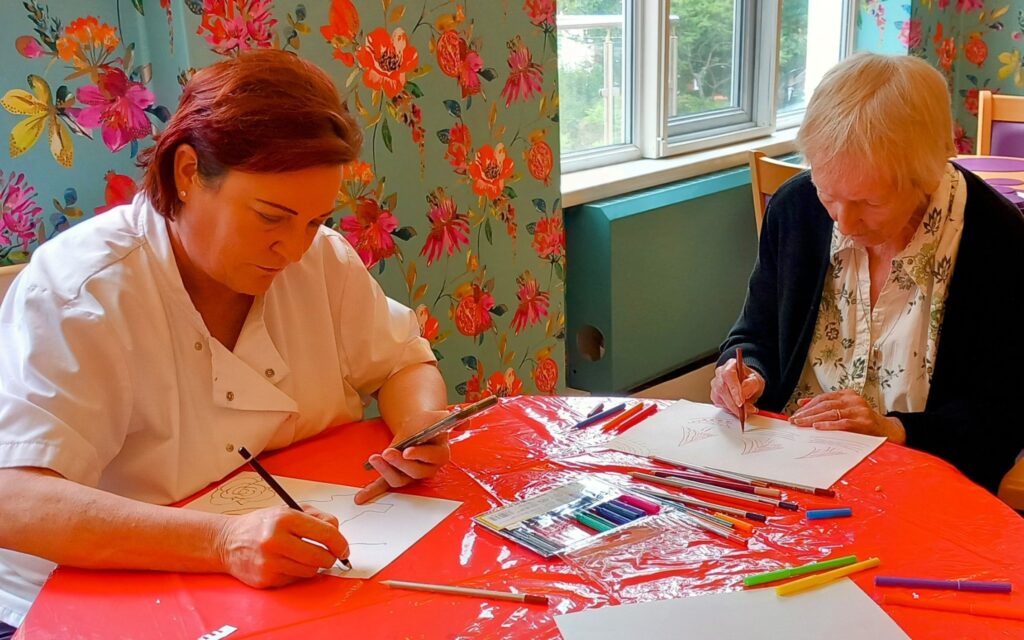 Two women sit at a table covered with red plastic, smiling as they draw on paper with coloured pencils and felt-tip pens in a brightly decorated room with floral wallpaper.
