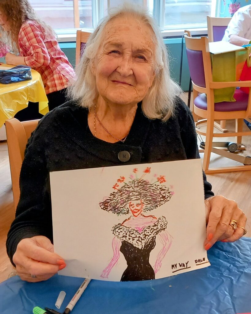An elderly woman sits at a table, smiling and holding up a colourful drawing of a woman in a black dress and large hat. Art supplies for sketching are scattered across the table.
