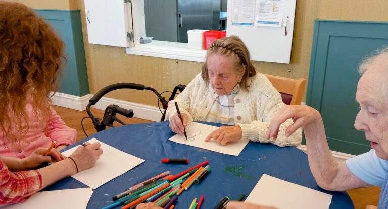 Three people are sitting at a table, smiling as they sketch on paper with felt-tip pens; art supplies are scattered across the table.