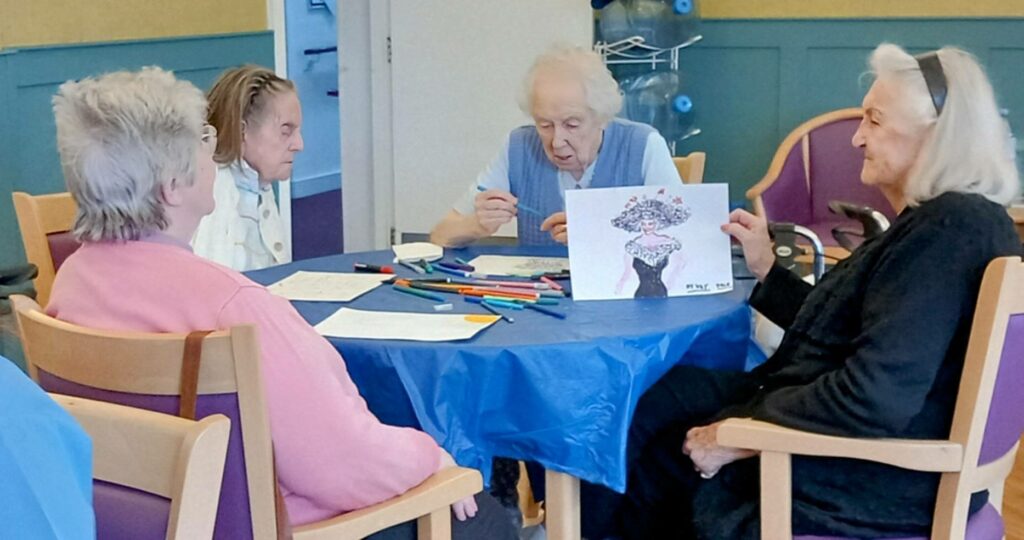 Four elderly women sit around a table, smiling as they engage in sketching and arts and crafts with coloured pencils and finished artwork in a communal room.