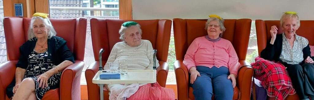 Four elderly women are seated in chairs, each with a coloured beanbag on her head, in a well-lit room with large windows in the background.
