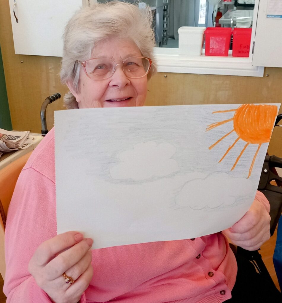An elderly woman with glasses sits indoors, smiling and holding up a drawing of clouds and the sun in the sky.