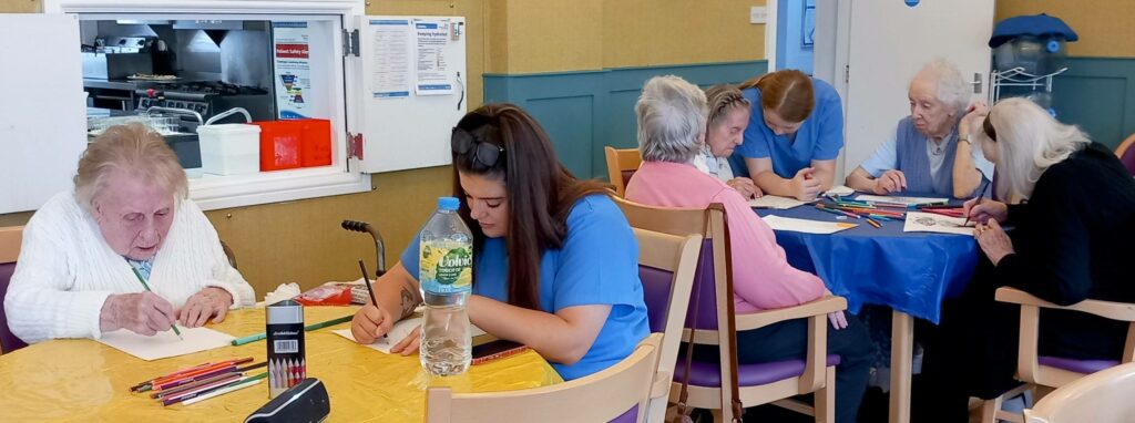 Elderly women and carers sit at tables in a care home, smiling as they enjoy sketching and colouring with coloured pencils and paper.