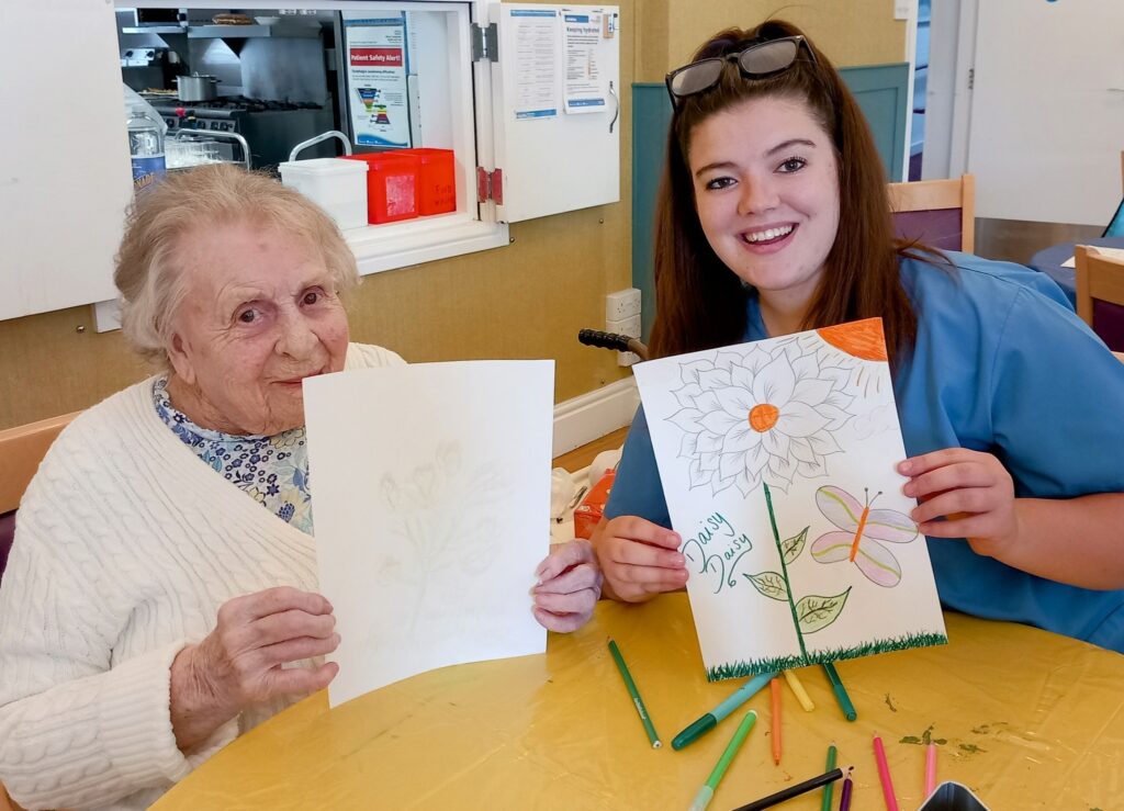 An elderly woman and a young woman sit at a table, smiling as they each hold up a drawing. Coloured pencils are spread out on the table in front of them, hinting at an afternoon spent sketching together.