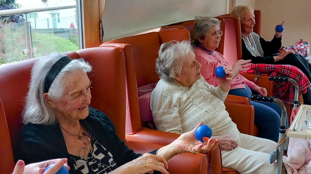 Four elderly women are seated in armchairs indoors, each holding a blue exercise ball in her right hand. They are enjoying Gentle Moves together and sharing good times during this group activity.