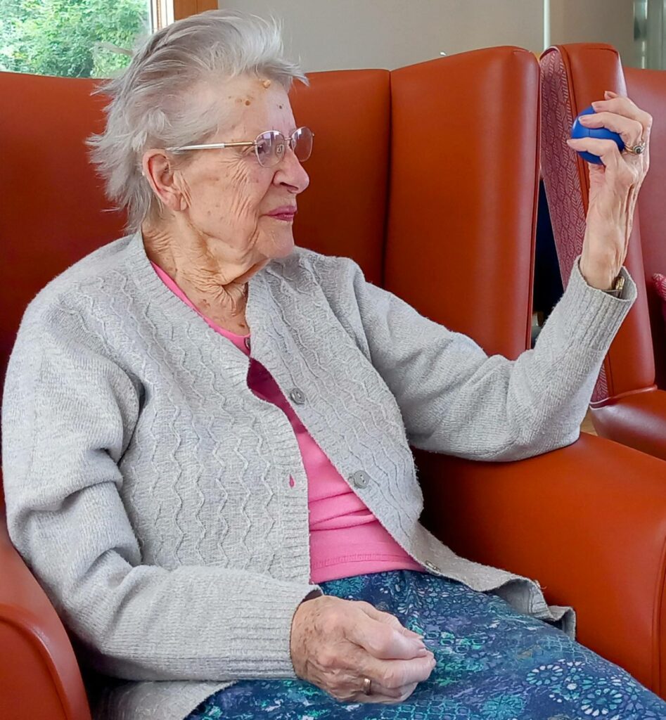 Elderly woman sitting on an orange chair, holding up a small blue object in her right hand and looking at it, enjoying Good Times and gentle movements in her cosy space.