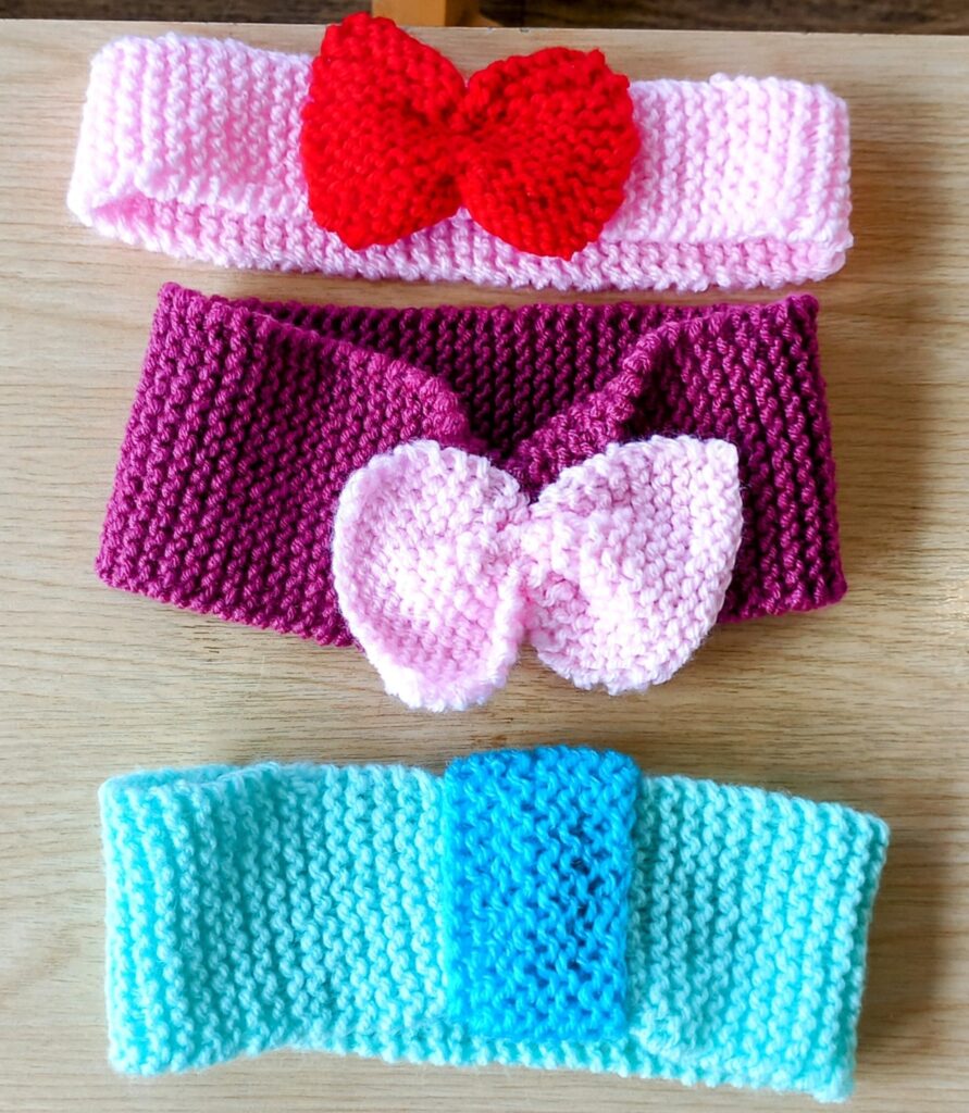 Three hand-knitted headbands on a wooden surface: one pink with a red bow, one purple with a pink bow, and one light blue with a blue accent.