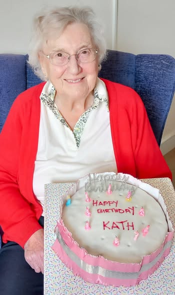 An elderly woman with white hair and glasses, wearing a red cardigan, sits and smiles while holding a birthday cake that says Happy Birthday Kath with pink candles.