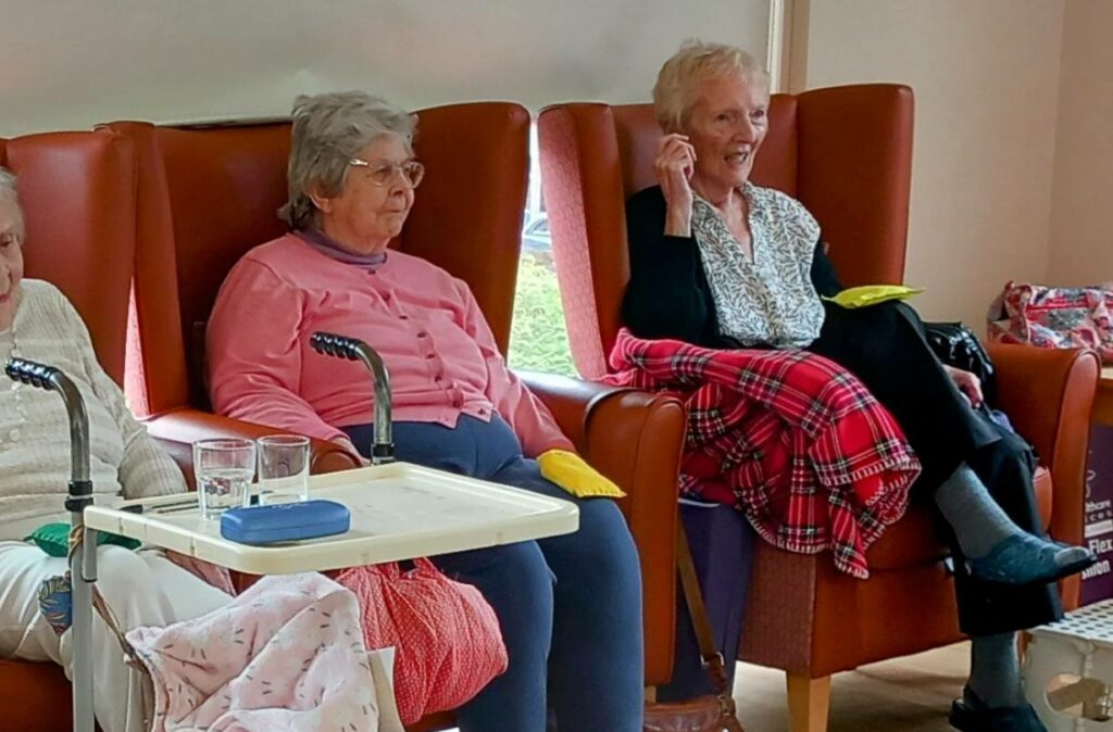 Three elderly women are seated on armchairs in a room, enjoying each other’s company. Walking aids and personal belongings are nearby, and one of the women has a blanket over her lap.