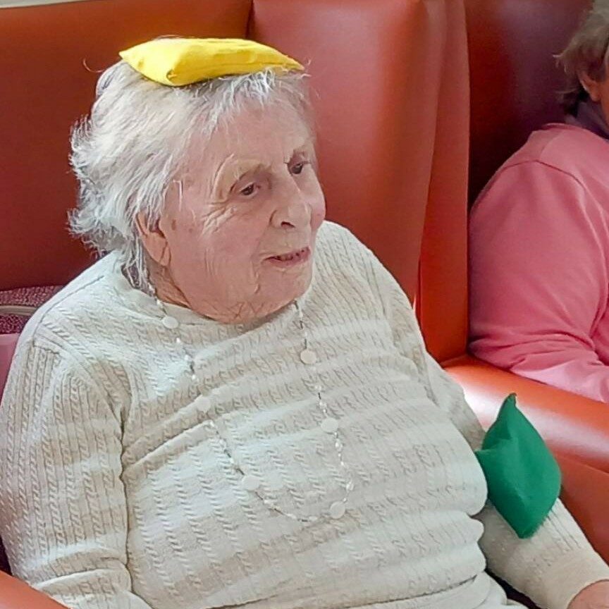 An elderly woman enjoys Good Times as she sits in a red chair, with coloured bean bags gently balanced on her head, arm, and lap.