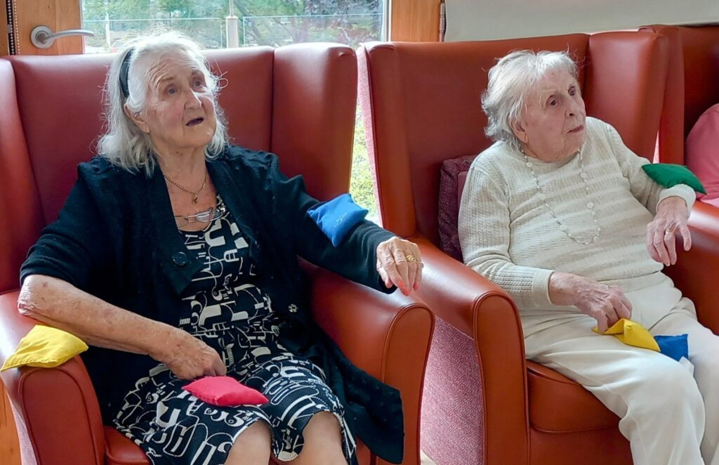 Two elderly women sit in red armchairs, each holding colourful beanbags on their laps and arms, enjoying Gentle Moves together and sharing Good Times in a well-lit room.
