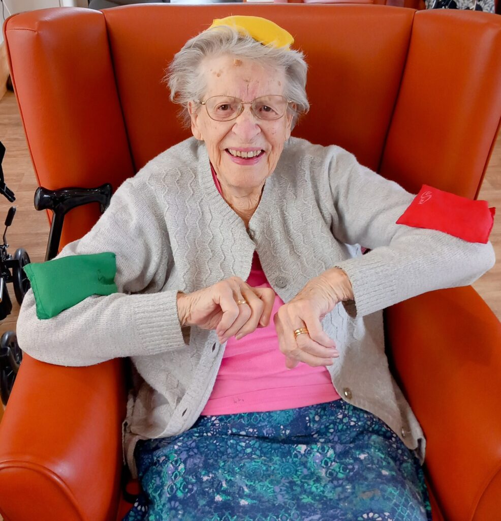 Elderly woman sitting in an orange armchair, smiling, with coloured beanbags on her head and arms, enjoying some good times and gentle movements.