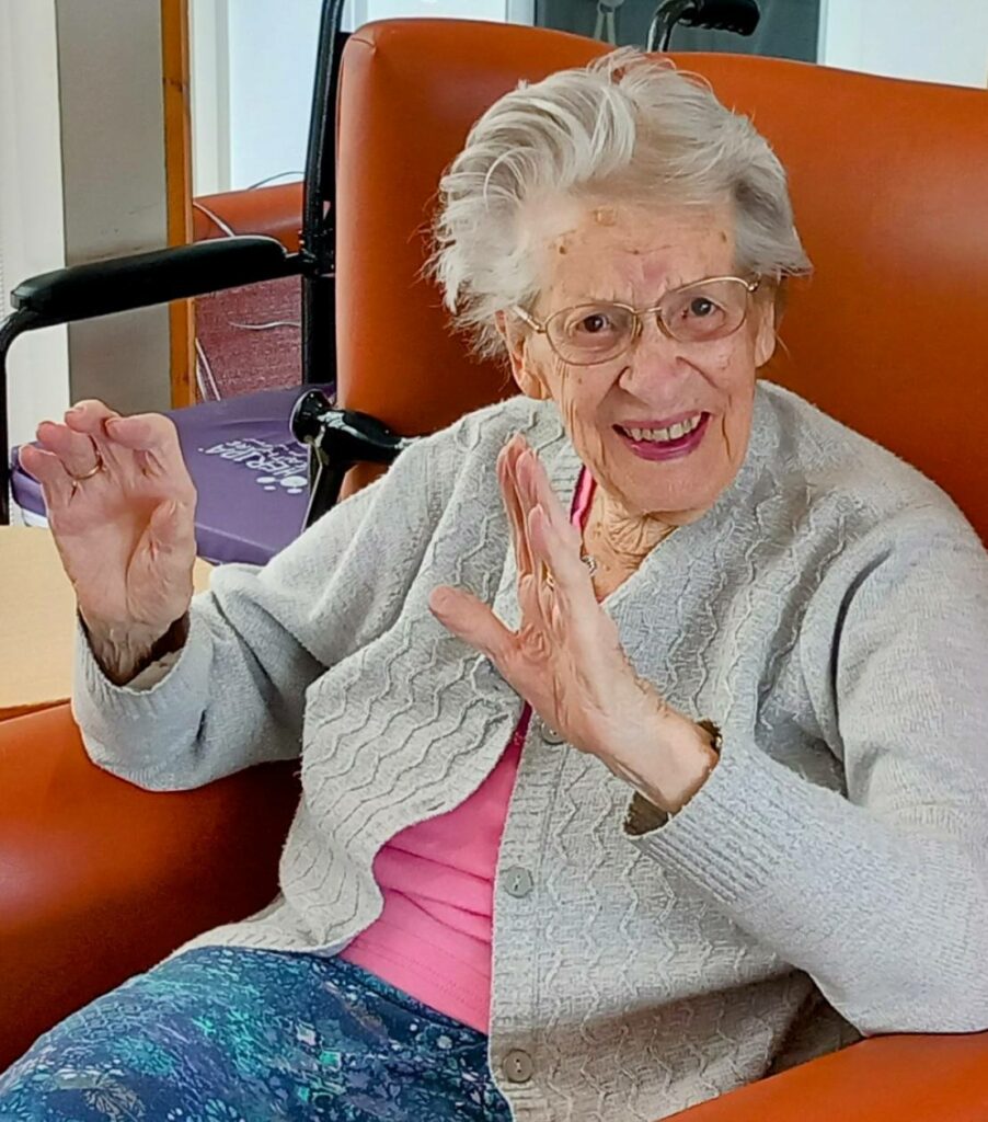 An elderly woman with grey hair, glasses, and a light cardigan sits in an orange chair, smiling and performing Gentle Moves with both hands raised, clearly enjoying the Good Times.