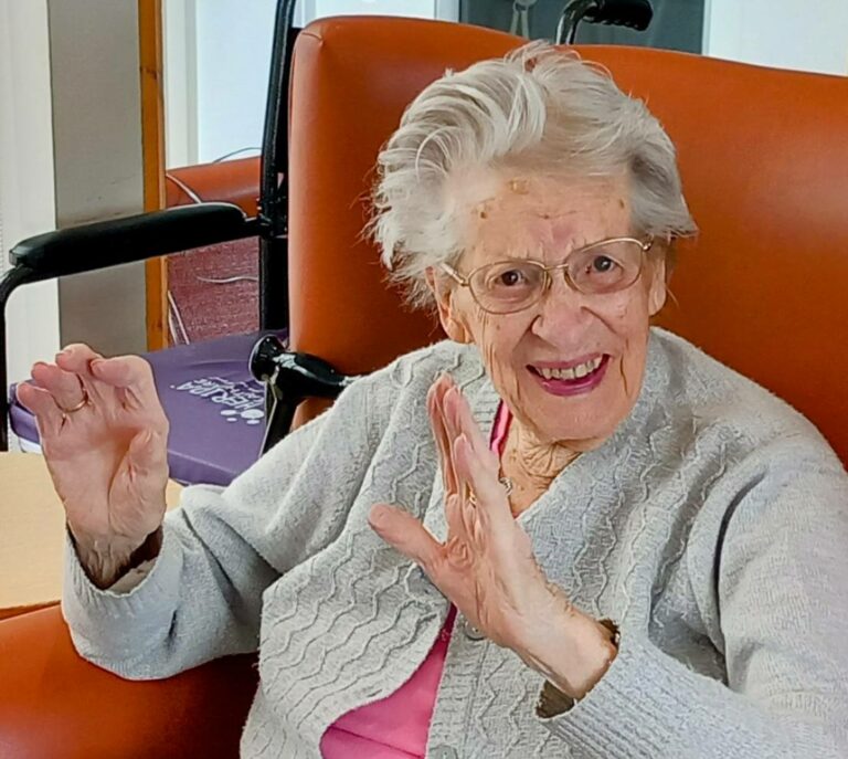 An elderly woman with grey hair, glasses, and a light cardigan sits in an orange chair, smiling and performing Gentle Moves with both hands raised, clearly enjoying the Good Times.