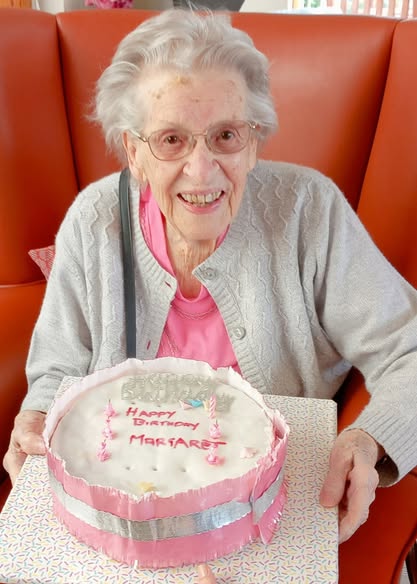 An elderly woman with spectacles smiles while holding a cake decorated with pink candles and the words “Happy Birthday Margaret,” celebrating Margaret’s birthday.
