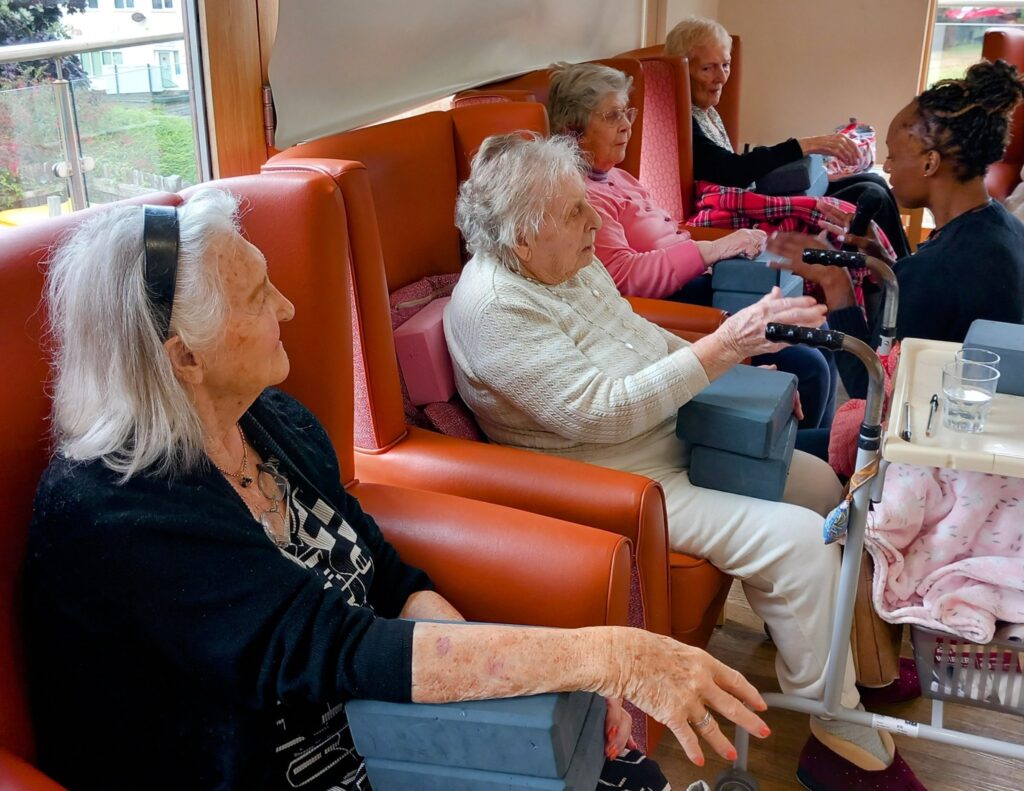 Elderly women seated in a row enjoy Good Times as carers provide manicures in a brightly lit room with large windows.