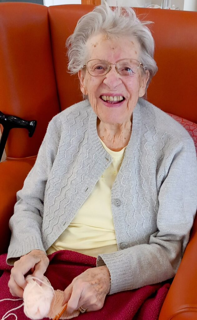An elderly woman with glasses sits in an orange chair, smiling, holding pink wool and knitting needles. She wears a light grey jumper and a yellow top. A red blanket covers her lap.