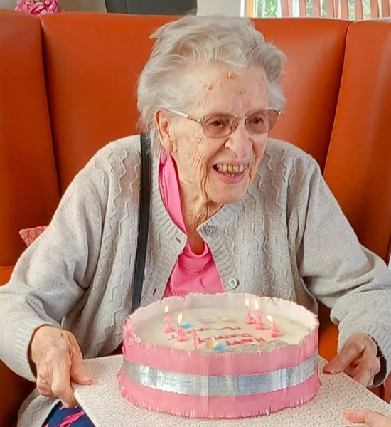 Elderly woman with grey hair and glasses smiles while holding a pink and white birthday cake with lit candles, celebrating Margaret’s birthday.