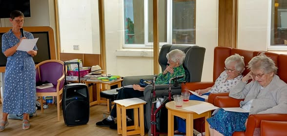 A woman stands reading from a sheet of paper while three elderly women sit and listen in a lounge area with tables, chairs, and papers.